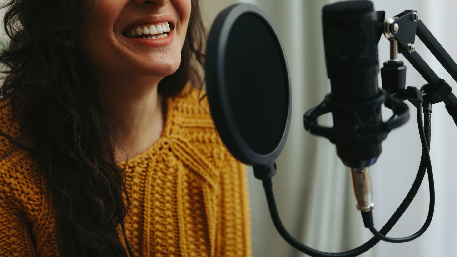 Woman standing in front of a microphone doing a voiceover. She is wearing an orange jumper and smiling 