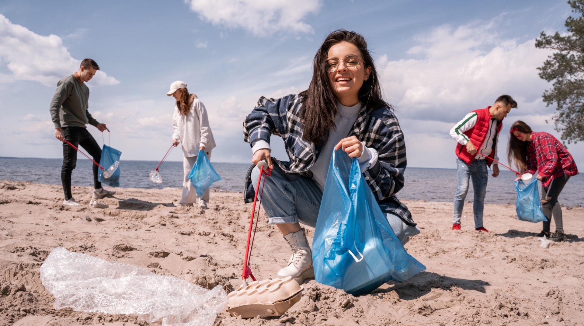 Woman picking up trash whilst being on a beach with other people also doing the same.