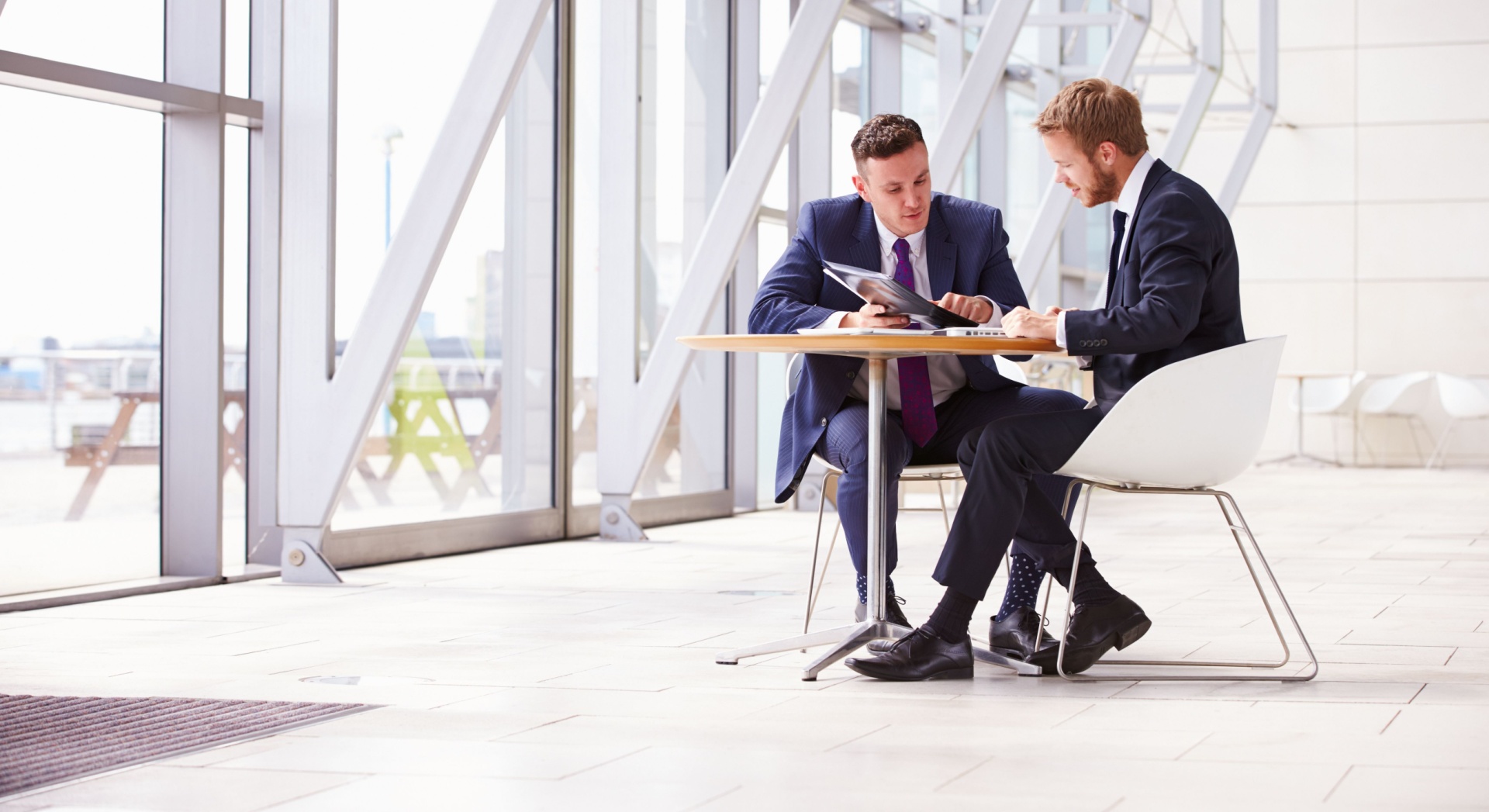 Two people dressed formally at a desk talking to each other in an empty white room 