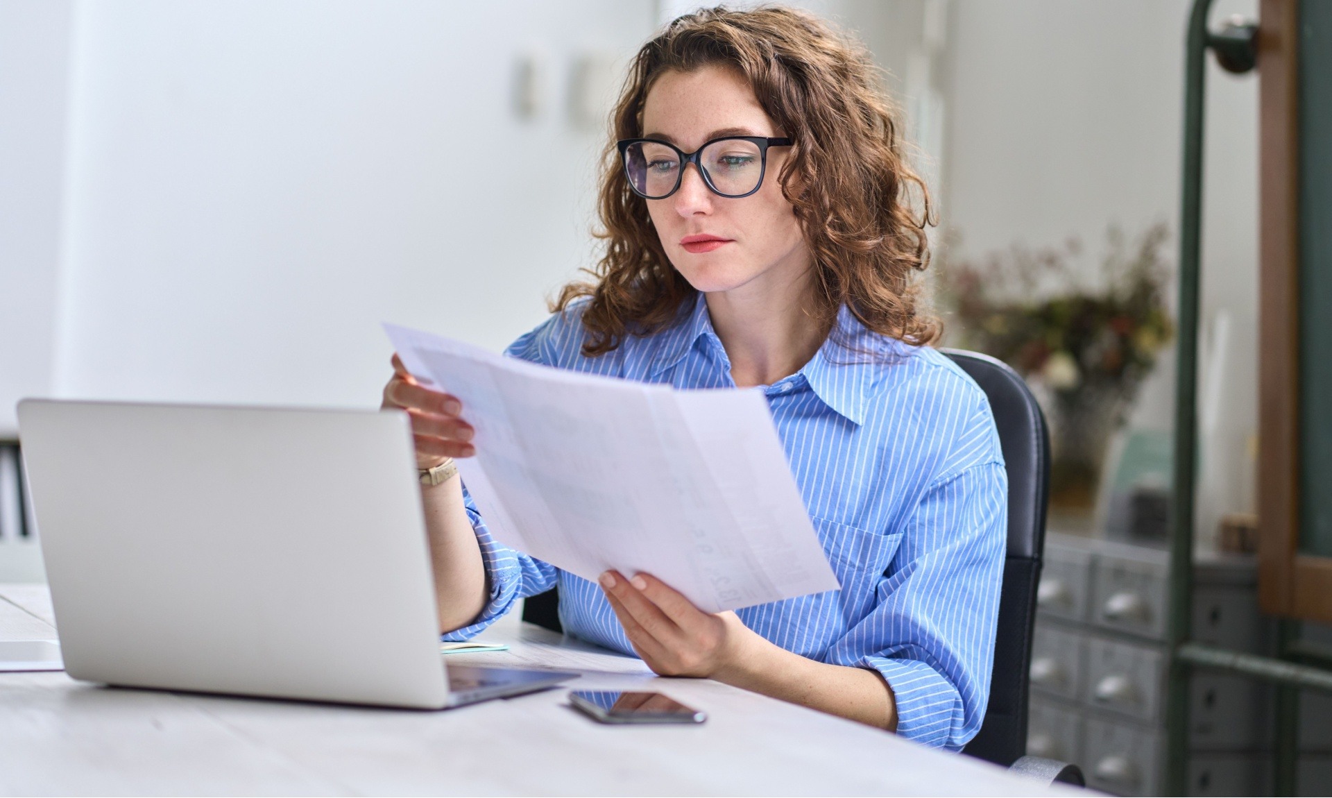 Woman doing her bookkeeping with an open laptop in front of her whilst holding a pen and paper. 